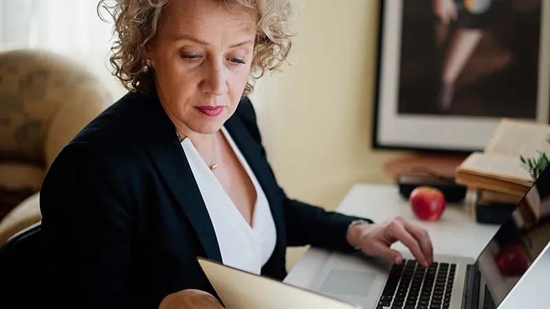 A person in a dark blazer working at a laptop with paper documents on the desk