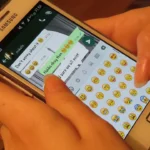 Hands holding a Samsung phone showing a WhatsApp chat with the emoji keyboard open