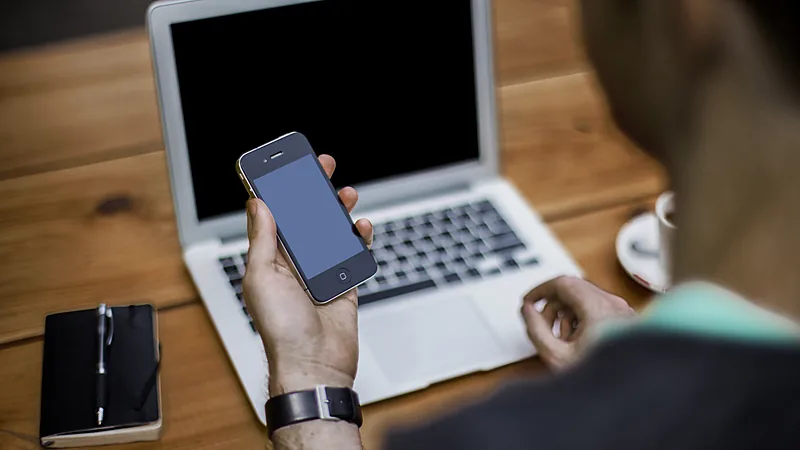 A man holding a smartphone next to an open laptop on a wooden desk
