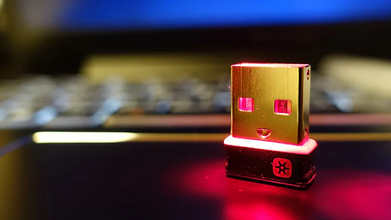 Close-up of a Logitech Unifying receiver standing upright with the orange Unifying logo visible and a keyboard blurred in the background