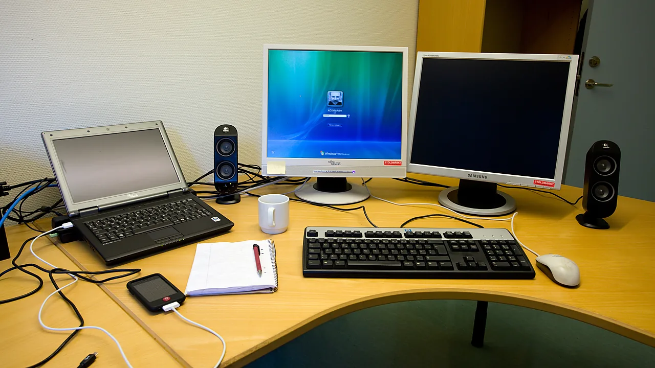 Office desk with a laptop, keyboard, and two monitors, one showing a Windows login screen