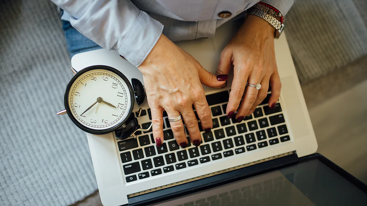 Hands typing on a laptop keyboard with an alarm clock beside the device