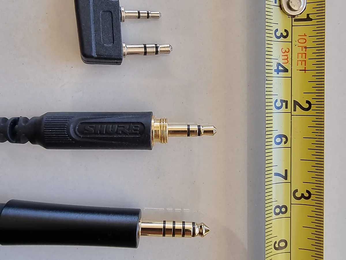 Several 3.5 mm audio plugs laid side by side next to a tape measure, with different numbers of black rings visible on each.