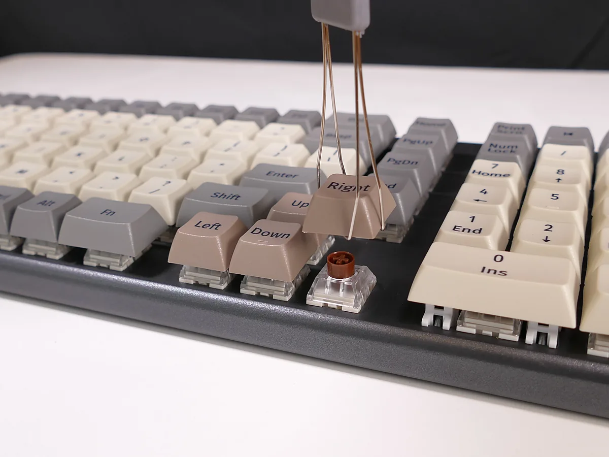 A keycap being lifted off a mechanical keyboard with a wire puller, exposing the switch underneath, with the arrow keys and number pad around it.