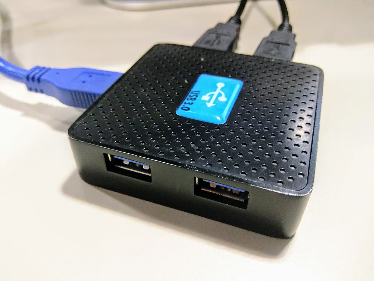 A small black four-port USB 3.0 hub with two cables plugged into it, sitting on a pale desk surface.